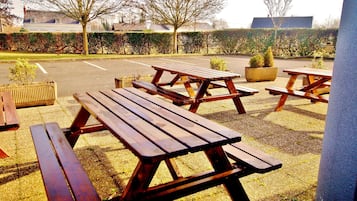 Outdoor picnic tables arranged in a row with a hedge and trees in the background.