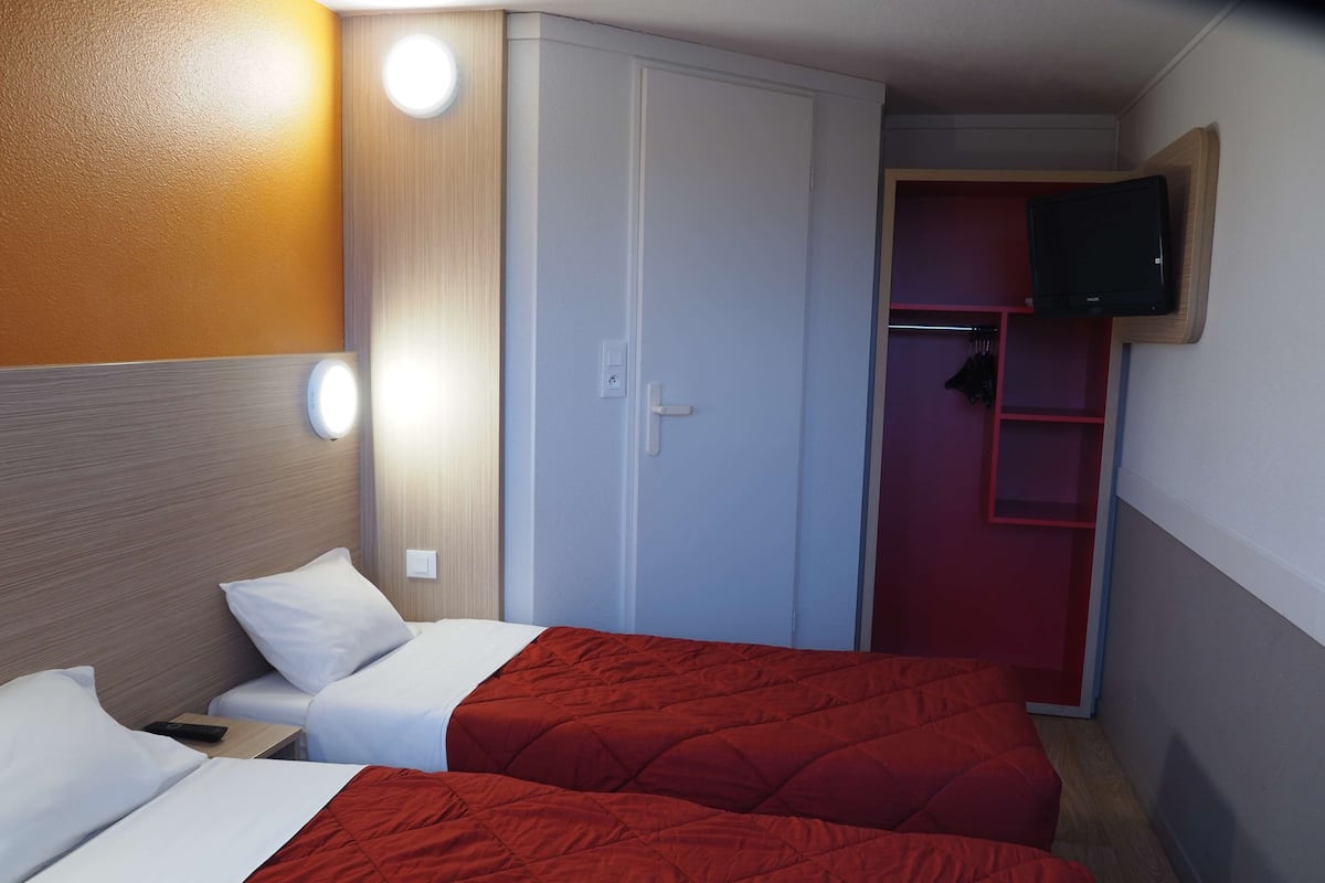 A hotel room with two beds, a wardrobe, a TV mounted on the wall, and a red storage unit.