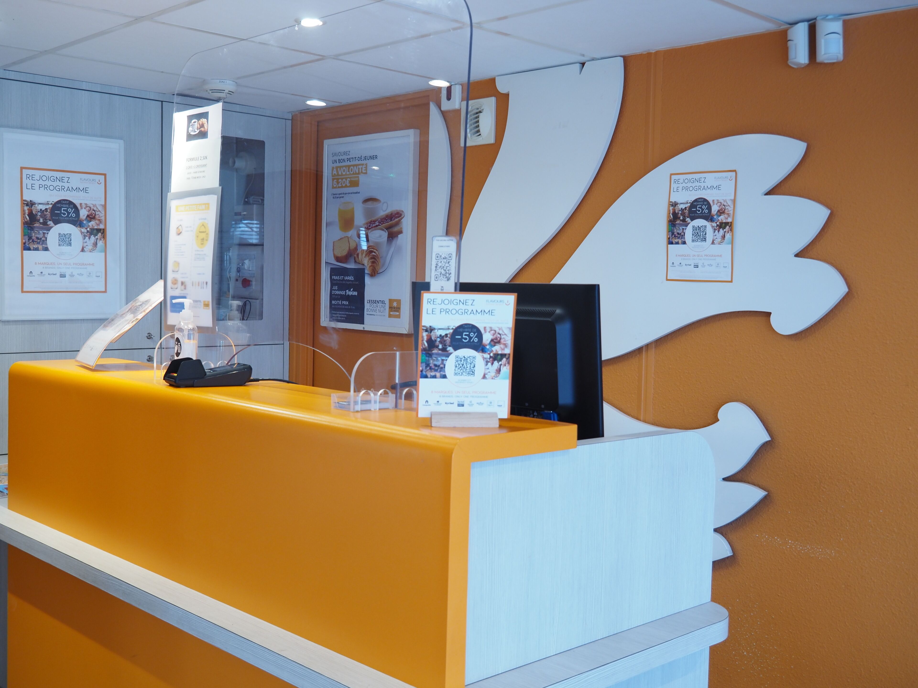 A reception desk with a yellow counter, a black phone, and a digital screen. There are framed posters on the wall and a large white decorative element.
