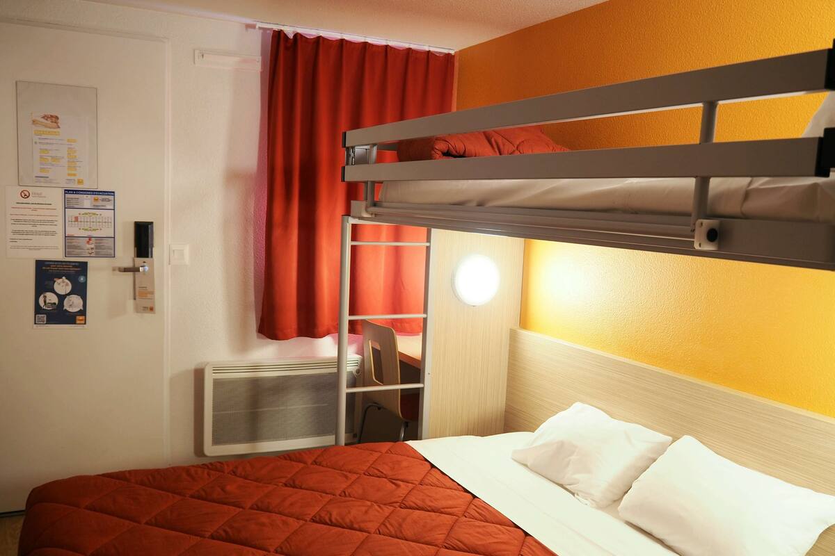 A bunk bed with a red quilted bedspread and white pillows, a desk with a chair, a heater, and a window with red curtains.