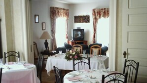 A dining area with round tables set for four, each with white tablecloths and floral centerpieces. There are two televisions on a wooden stand, and the room has a traditional decor with floral curtains. - Four Creeks Bed & Breakfast (Girard)