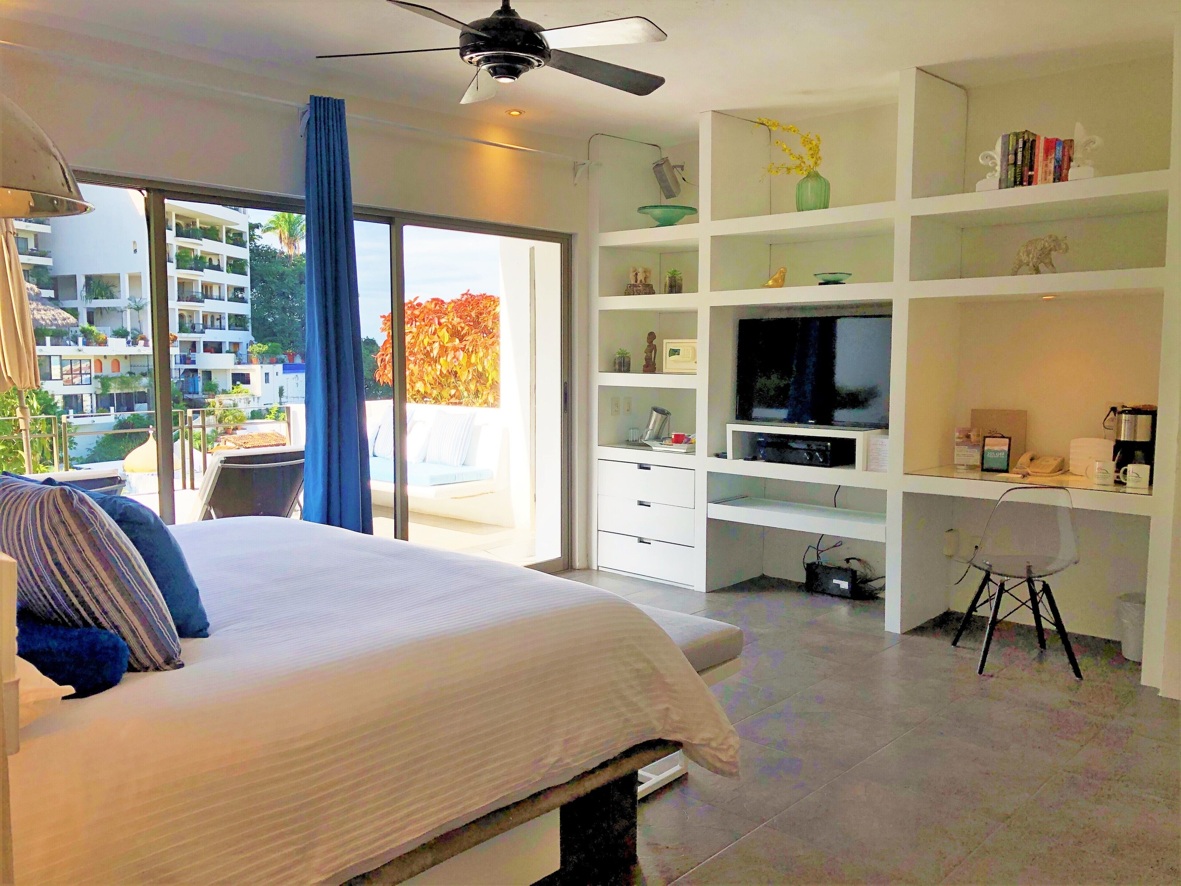Un dormitorio con cama, televisor, escritorio y balcón con vistas a edificios.
