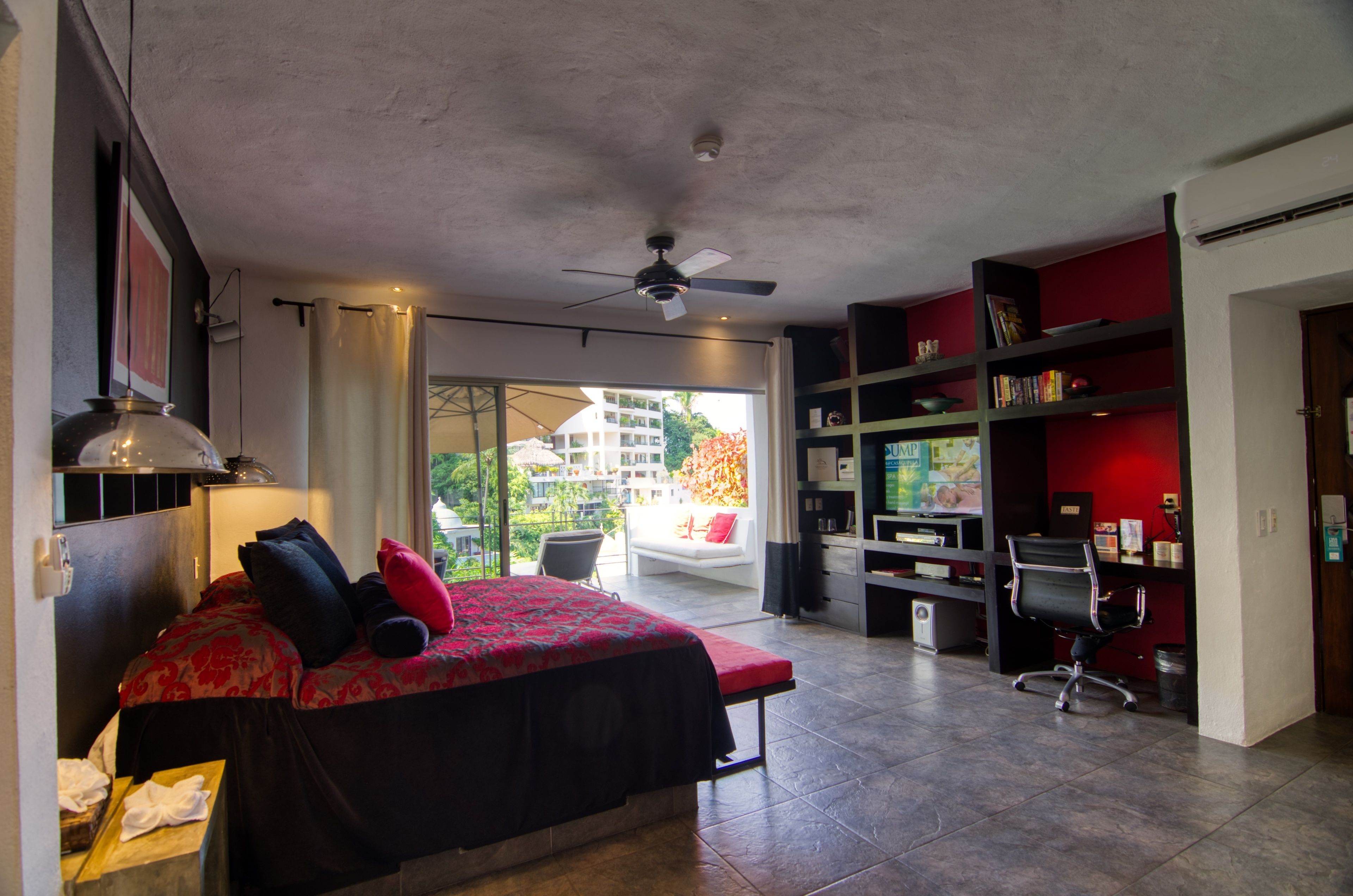Un dormitorio con cama, escritorio, silla, estantería y vistas al exterior.