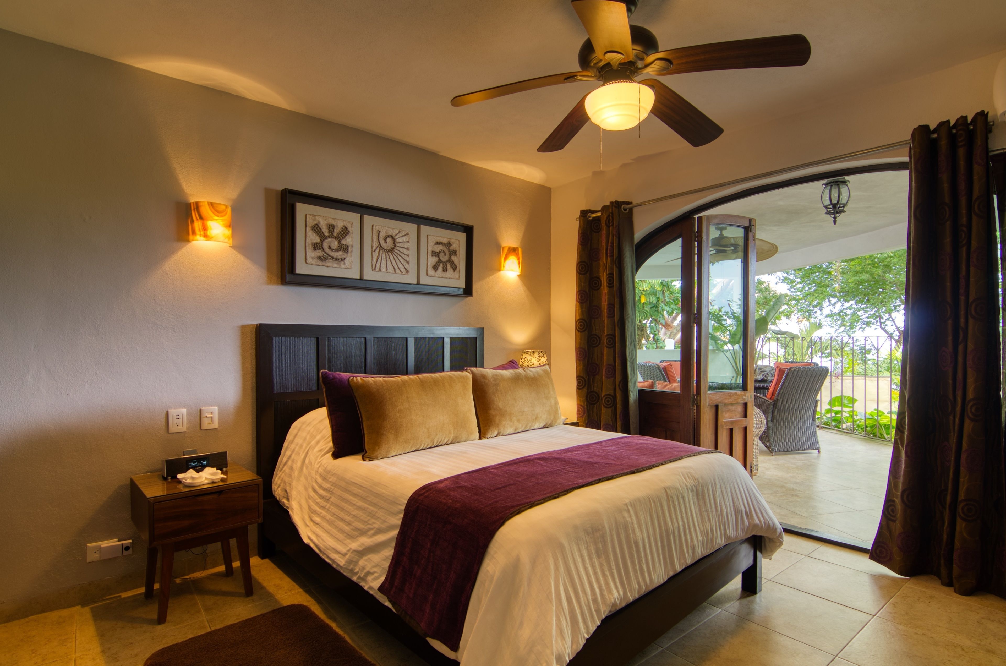 Un dormitorio con cama, ventilador de techo, mesita de noche y vistas al exterior.