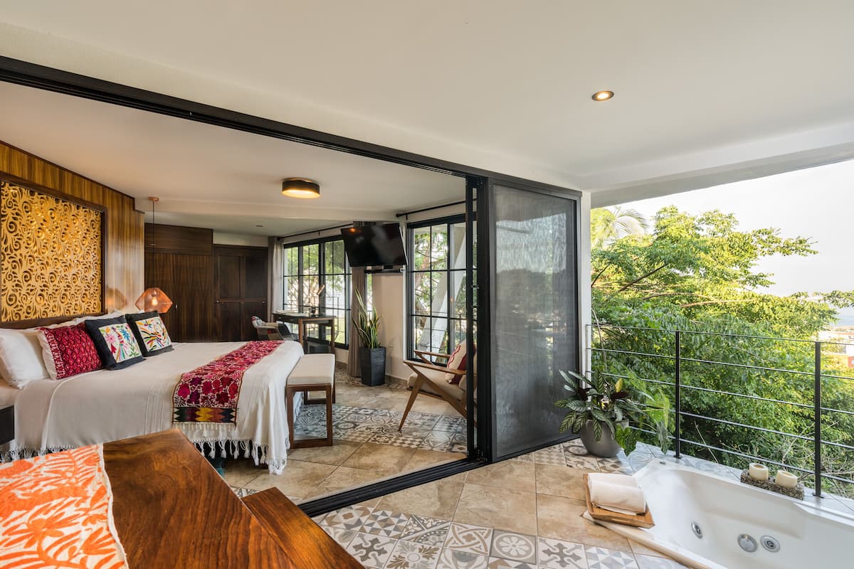 Un dormitorio moderno con una cama grande, un área de descanso y un balcón con vistas a zonas verdes.