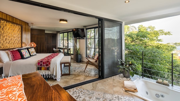 A modern bedroom with a large bed, a sitting area, and a balcony with a view of greenery.