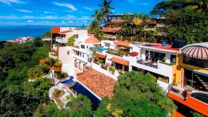 A modern multi-story building with a rooftop pool, surrounded by lush greenery and a view of the ocean.