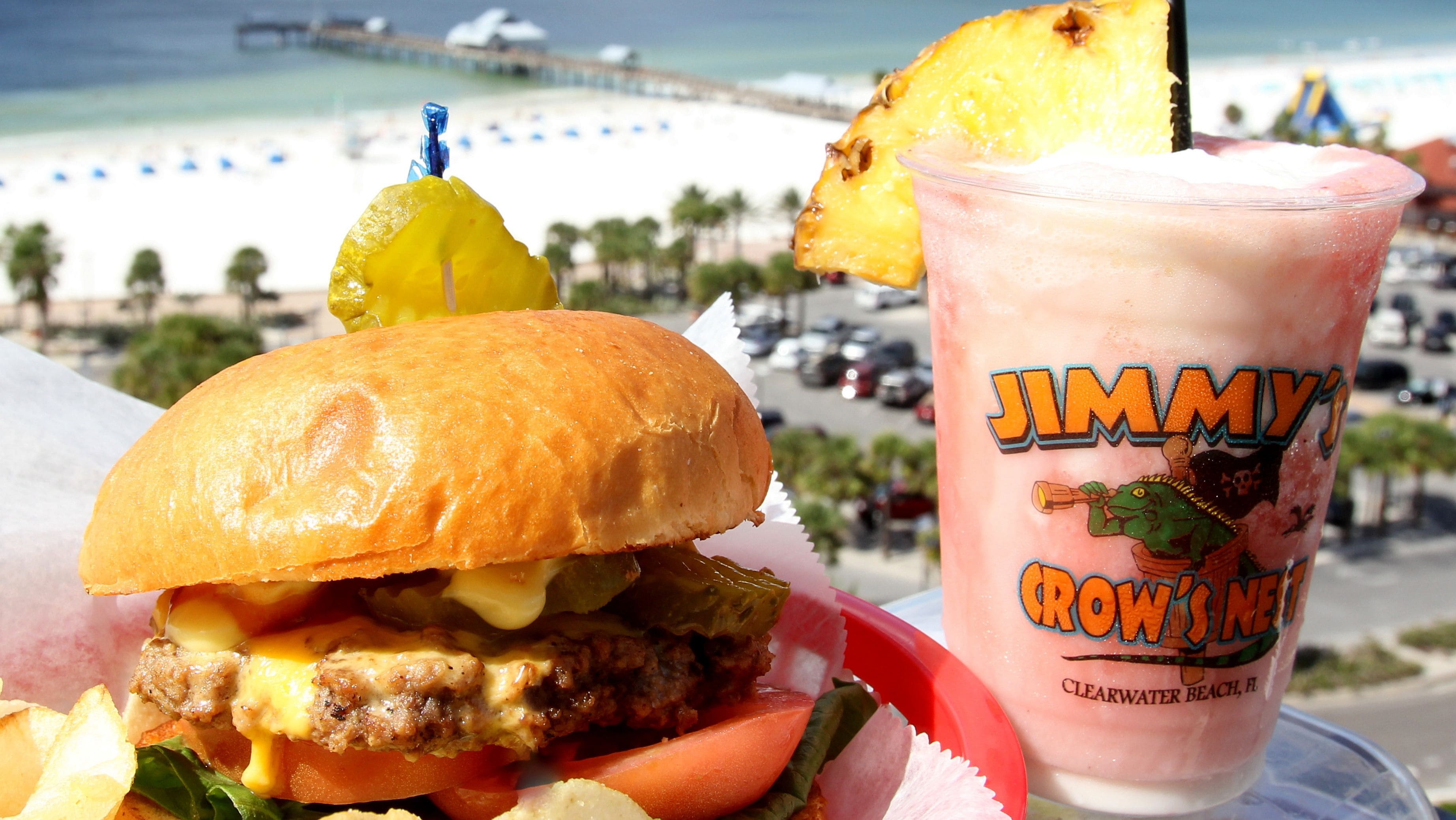 Ein Burger mit Ananas, ein Getränk mit der Aufschrift "Jimmy's" und im Hintergrund ein Strandblick.