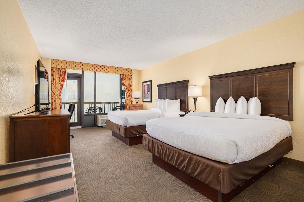 A hotel room with two beds, a desk, a chair, a window with curtains, and a view of the outside balcony.