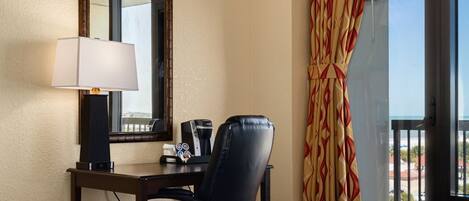 Habitación de hotel con cama, escritorio con lámpara, una silla, cafetera y ventana con cortinas.