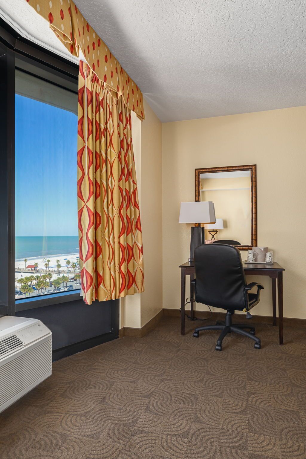 Een hotelkamer met een groot raam, een bureau met stoel, een lamp en een spiegel.