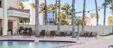 Zona de piscina con sillones reclinables, palmeras y un edificio con balcones.