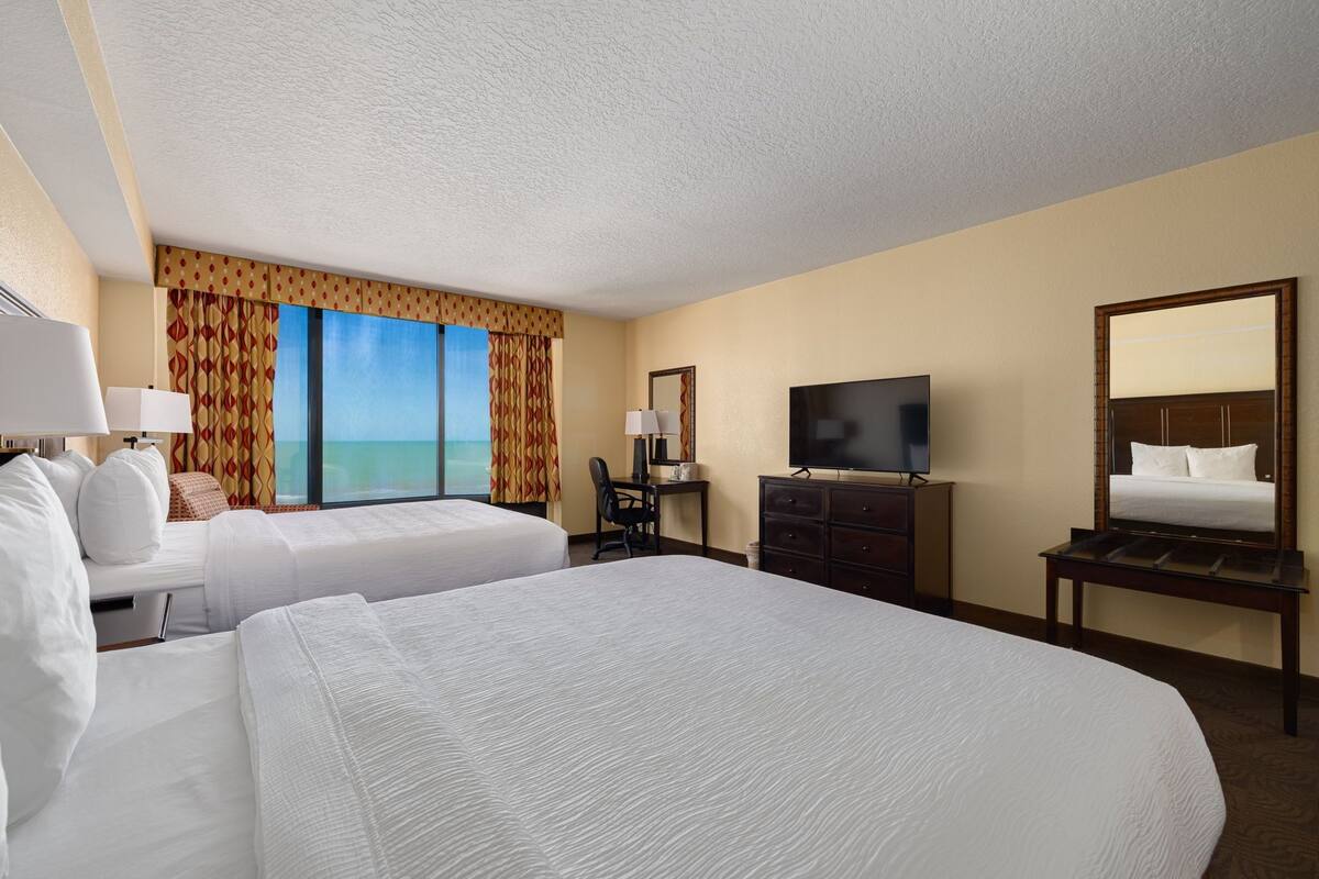 A hotel room with two beds, a desk, a TV, and a large window with curtains.