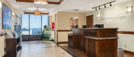 A reception area with a wooden reception desk, a large window offering a view of the beach, and a sign welcoming guests to the Pier 60 Marina Hotel.