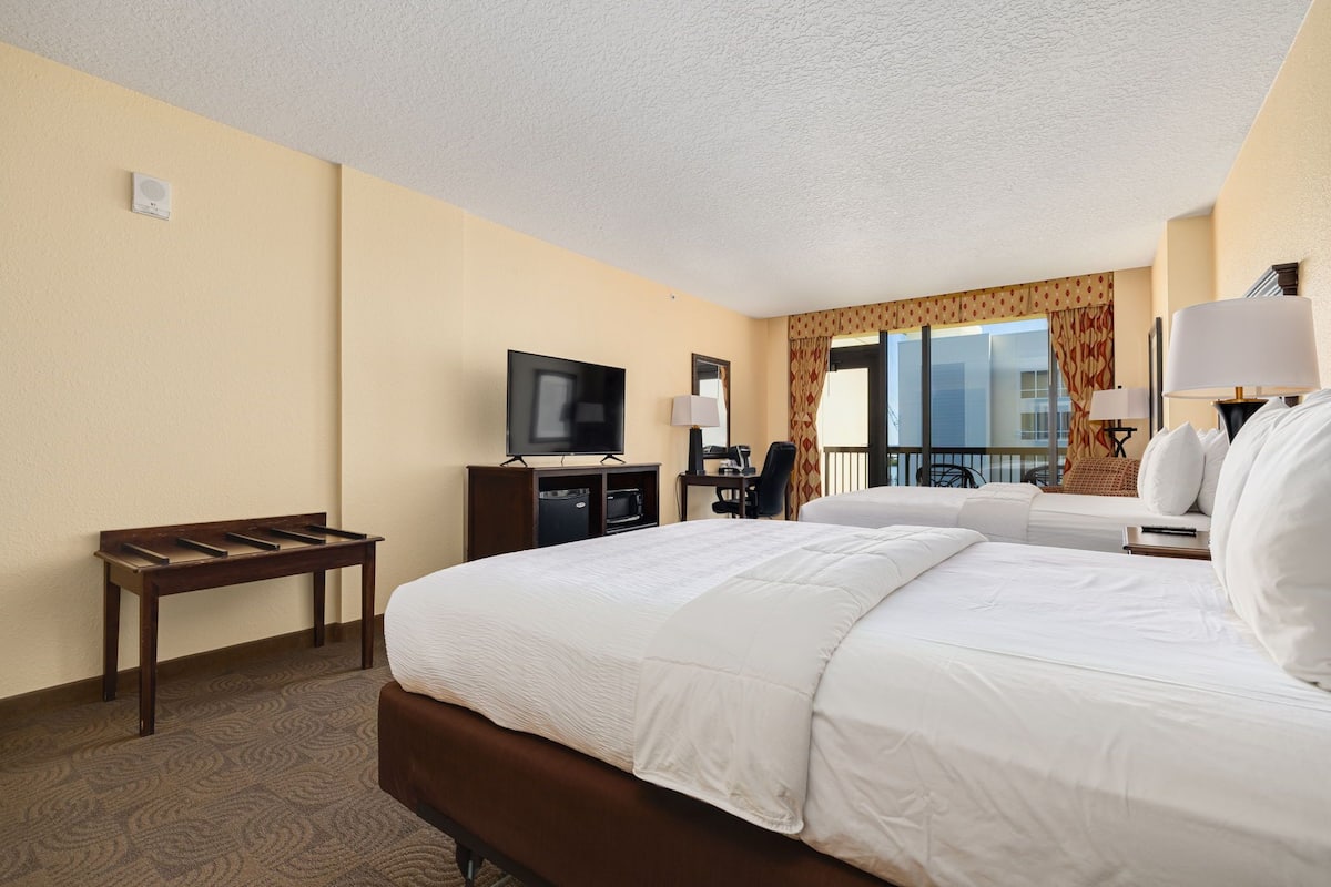 A hotel room with a large bed, a desk with a TV, a chair, and a window with a view.