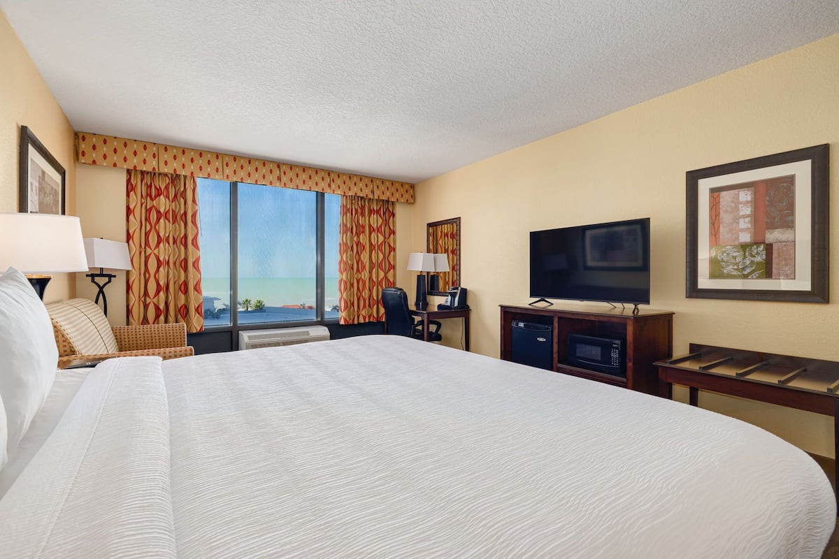 A hotel room with a large bed, a desk with a computer, a television, and a window with curtains.