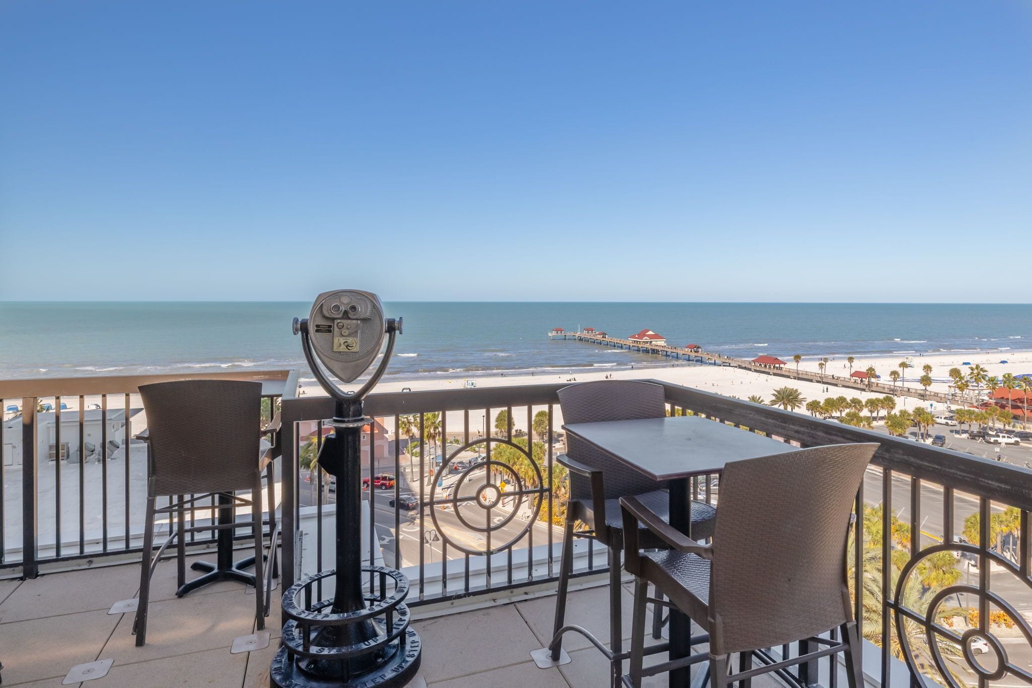 A balcony with a telescope, chairs, and a table overlooking a beach and pier.
