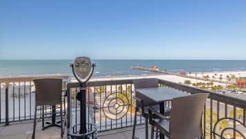 A balcony with a telescope, chairs, and a table overlooking a beach and pier.
