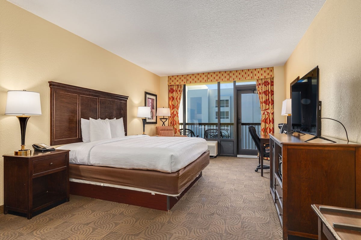 A hotel room with a large bed, a desk with a computer, a TV, and a balcony with curtains.