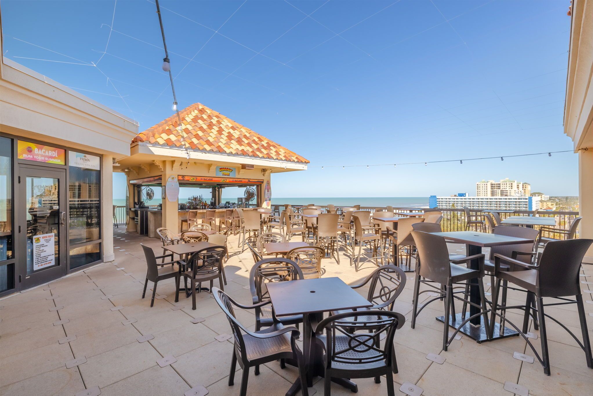 An outdoor patio area with tables and chairs, a tiled roof structure, and a view of the sea.