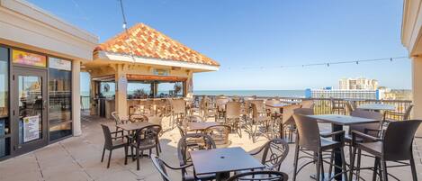 An outdoor patio area with tables and chairs, a tiled roof structure, and a view of the sea.