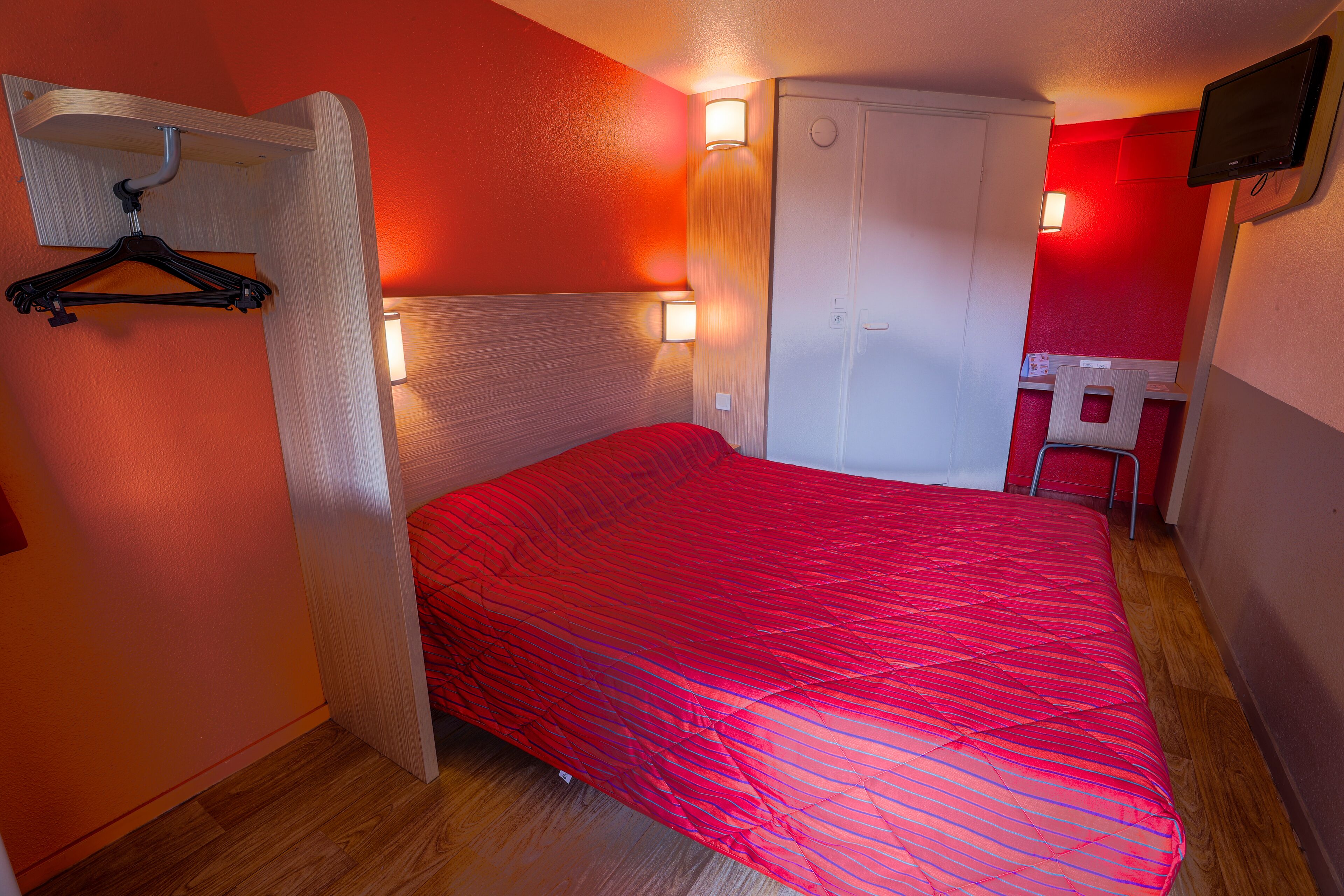 Een kamer met een bed, een houten hoofdeinde, een kapstok, een bureau met stoel en een aan de muur bevestigde televisie.