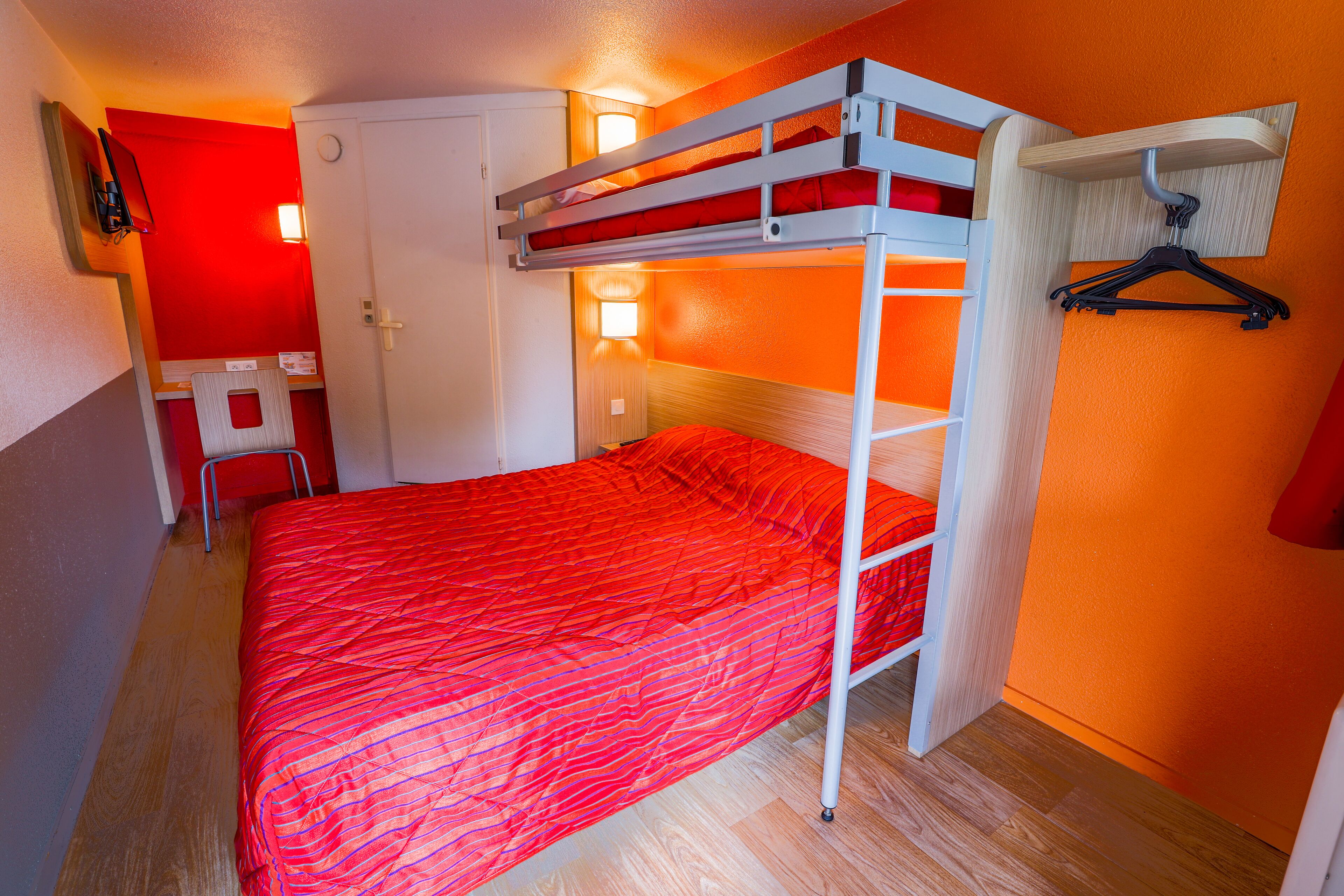 A bunk bed with a red bedspread, a desk with a chair, and a wall-mounted TV.