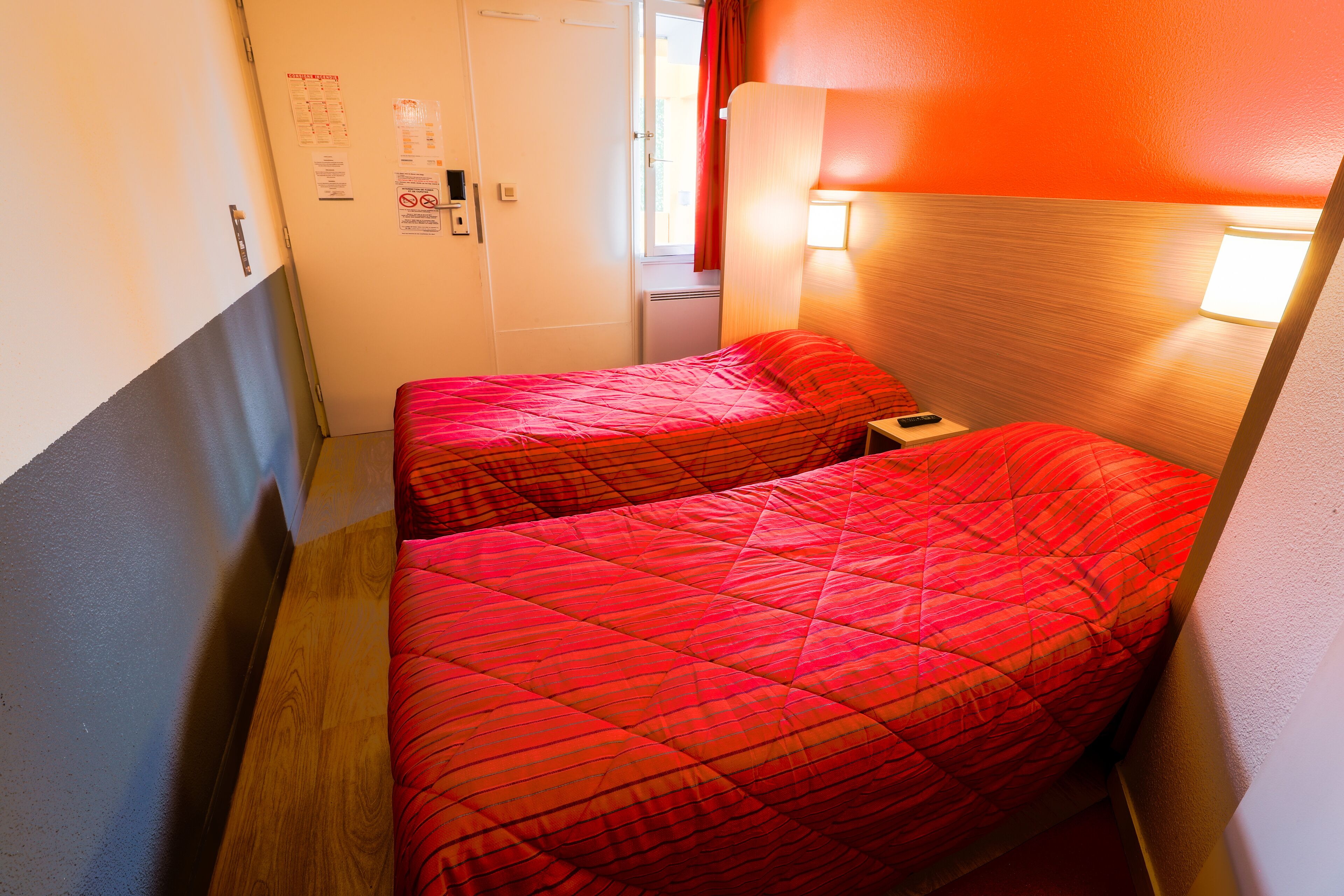 Une chambre d’hôtel étroite, avec deux lits, un plancher en bois et une porte donnant sur une salle de bain.