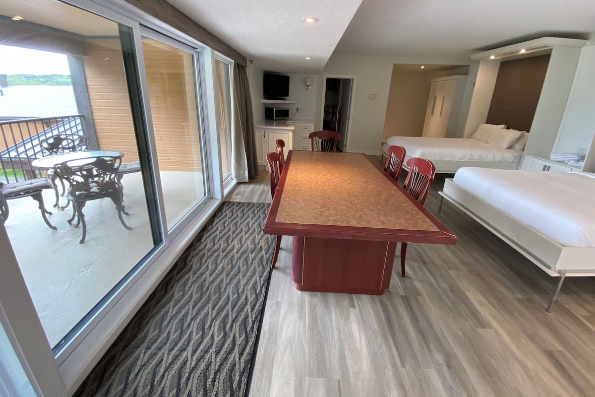 Quarto de hotel com uma grande porta de vidro deslizante que dá para uma varanda, piso de madeira, mesa de jantar central com cadeiras, cama e televisão em um móvel.