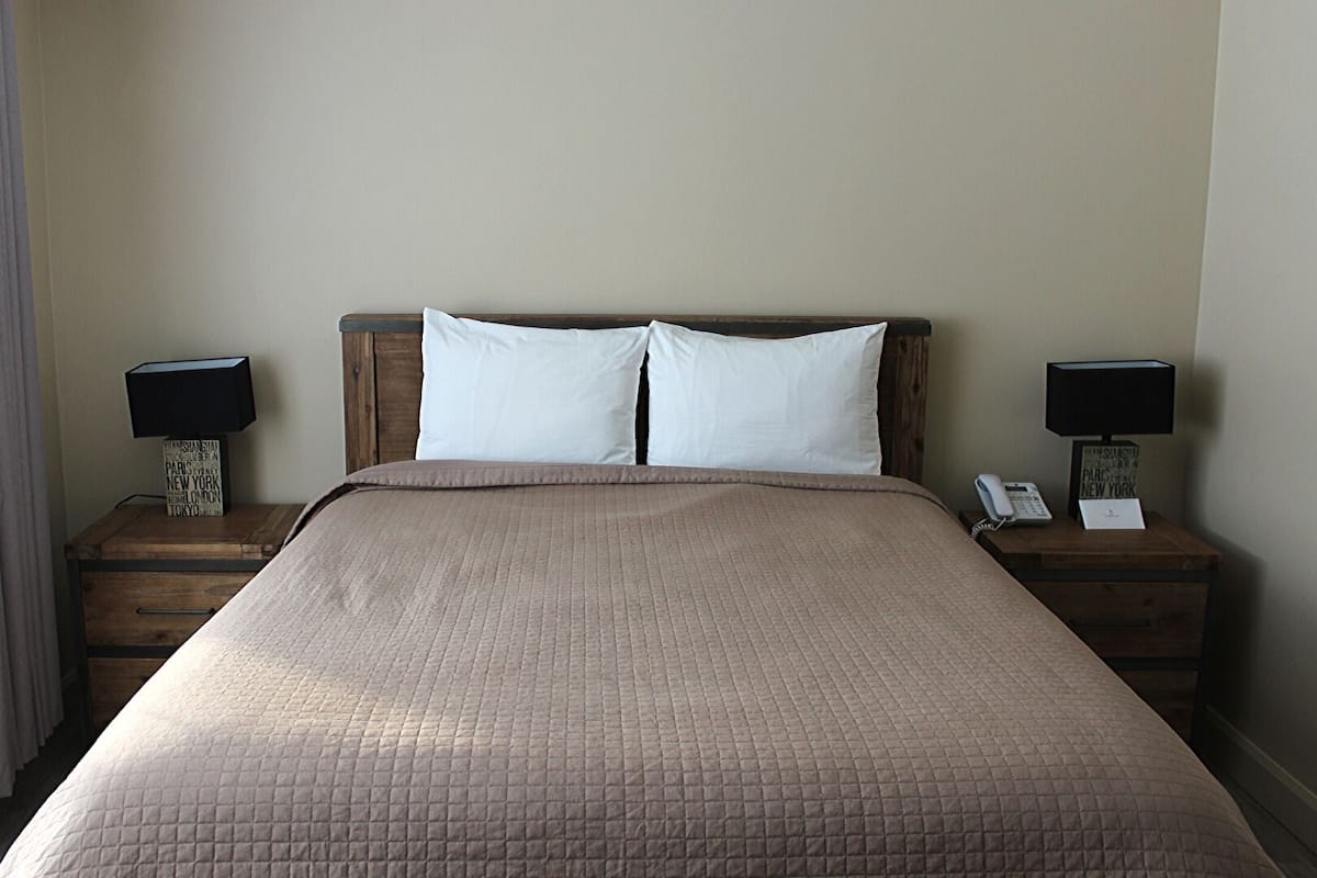 Uma cama bem arrumada com um edredom acolchoado, duas almofadas brancas e uma cabeceira de madeira. Duas mesas de cabeceira, com abajures e um telefone em uma delas.