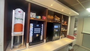 Un espace café en libre-service équipé d’une machine à café, d’un réfrigérateur et d’un présentoir où sont exposés divers produits alimentaires.