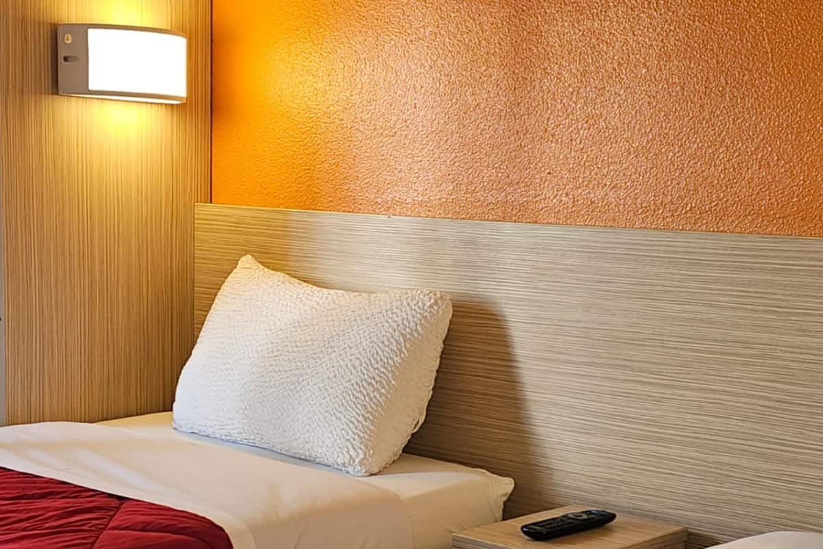 A hotel room with a wooden headboard, a white pillow, a red bedspread, and a remote control on a wooden nightstand.