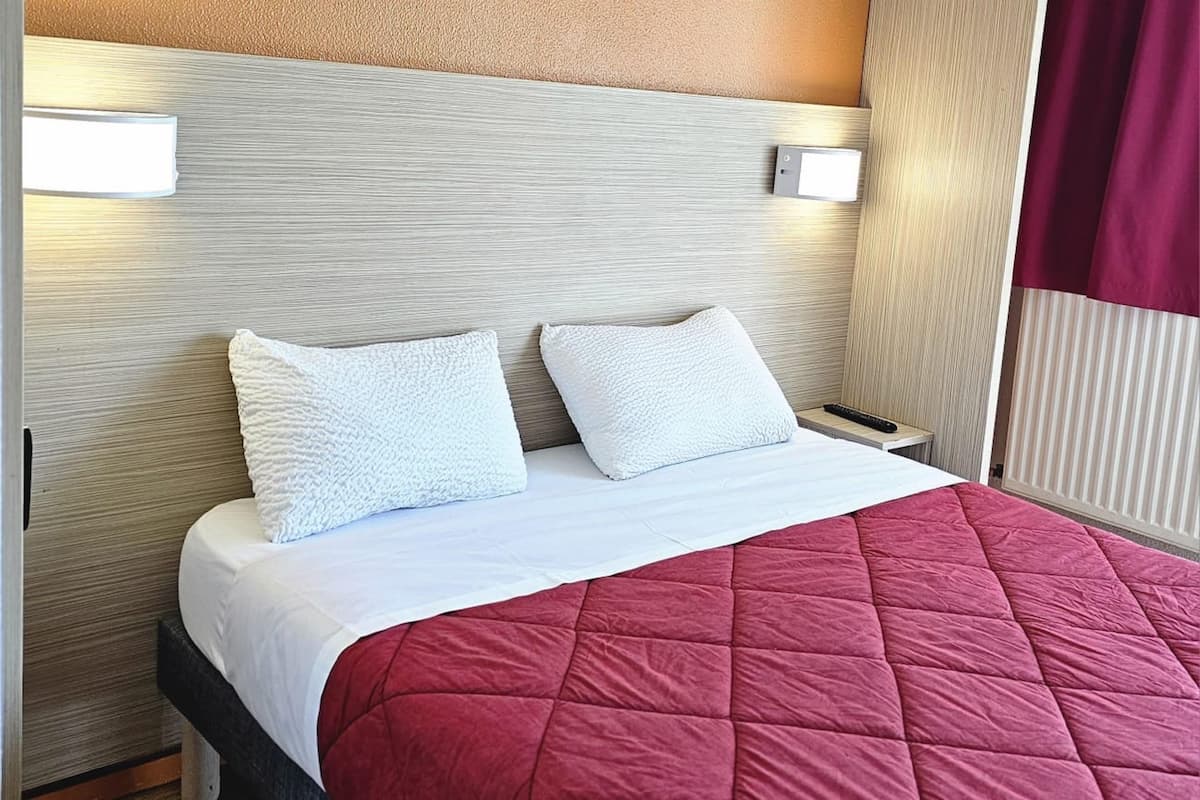 A neatly made bed with a red quilt, two white pillows, and a wooden headboard with wall-mounted lights.