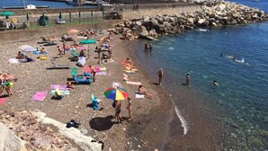 A beach with people sunbathing and swimming, a rocky breakwater, and boats in the background.
