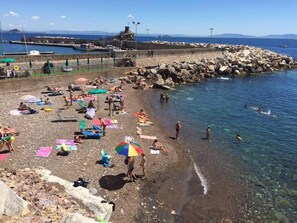 A beach with people sunbathing and swimming, a rocky breakwater, and boats in the background.
