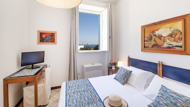 A hotel room with a bed, a desk with a computer, a window with a view, and artwork on the walls.