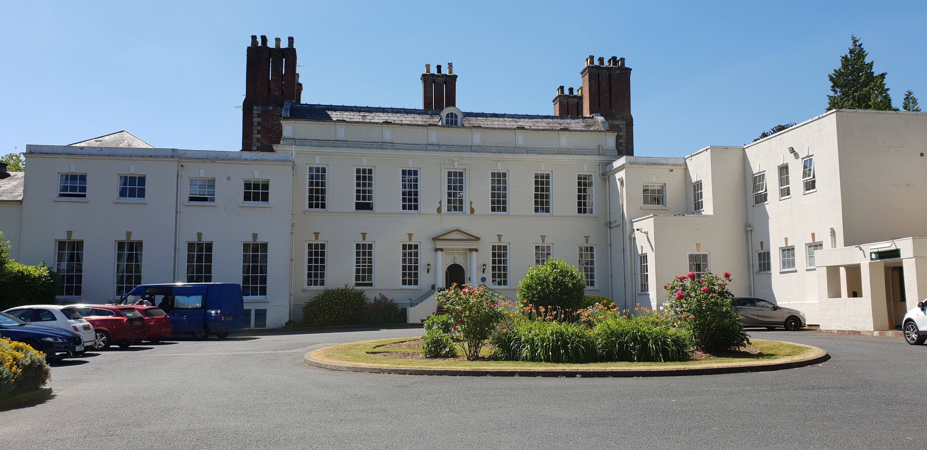A large, historic building with a symmetrical facade, multiple chimneys, and a central entrance. There are several parked cars and a blue van in the foreground.