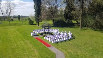 Disposición circular de sillas blancas con cintas moradas, una alfombra roja y un pabellón en un entorno al aire libre con césped.