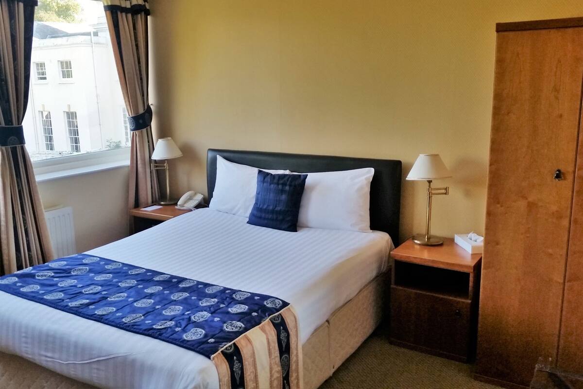 Habitación de hotel con una cama grande, una mesita de noche, una lámpara de lectura, un armario y una ventana con cortinas.