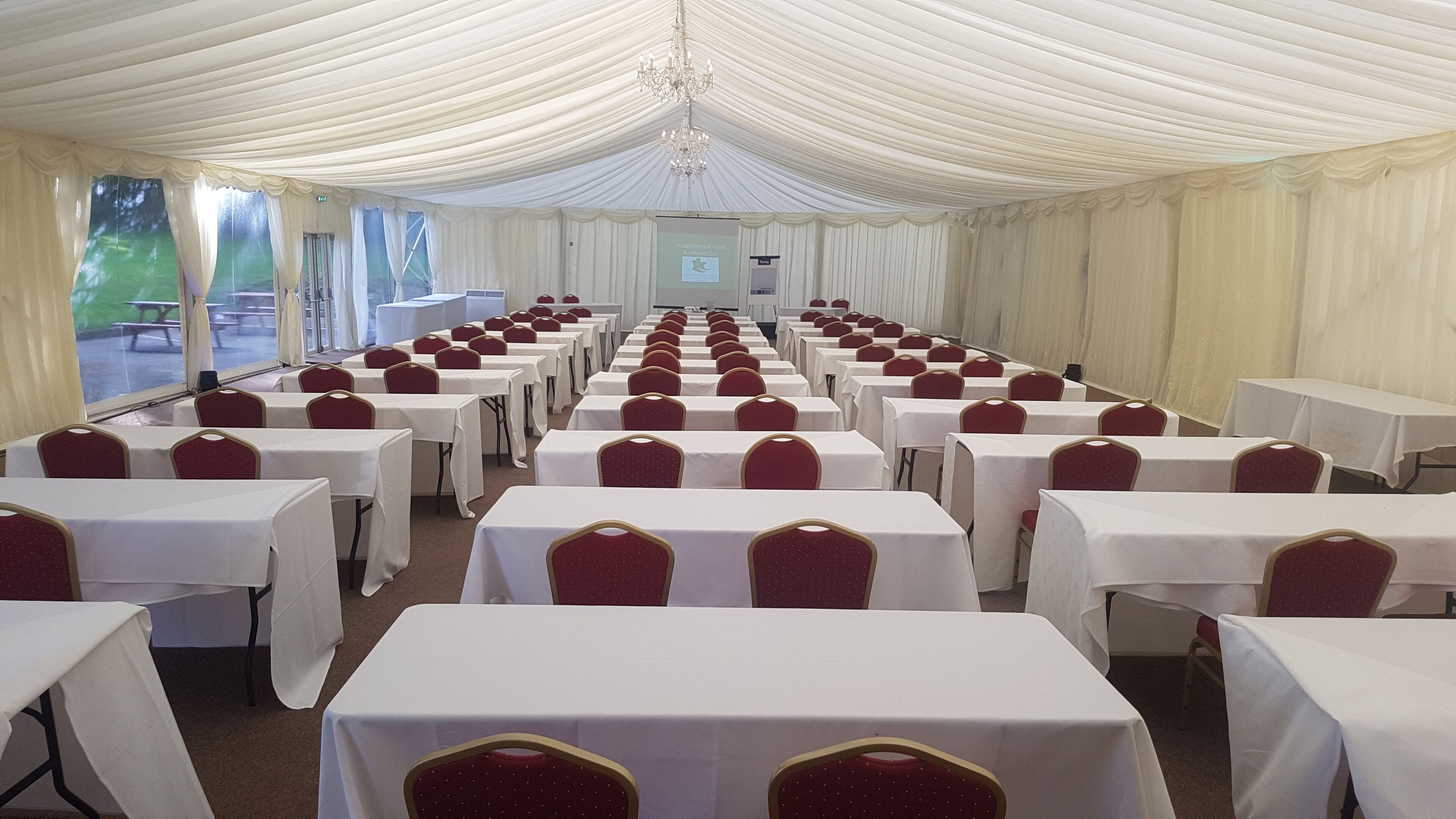A banquet hall with rows of tables and chairs, a chandelier, and a projection screen.