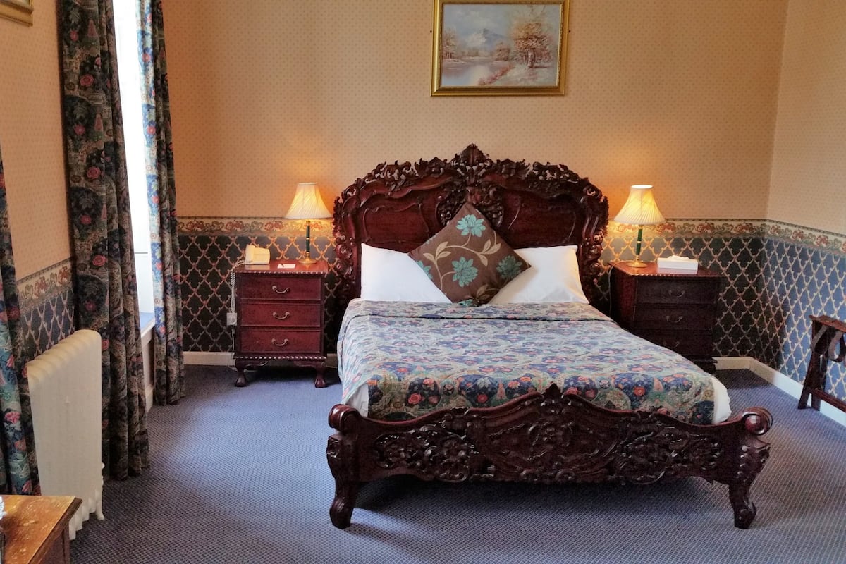 Un dormitorio con una cama grande, dos mesitas de noche con lámparas, un cabecero de madera, ropa de cama estampada con motivos florales, una mesita con una botella de agua y fruta, y un cuadro en la pared.