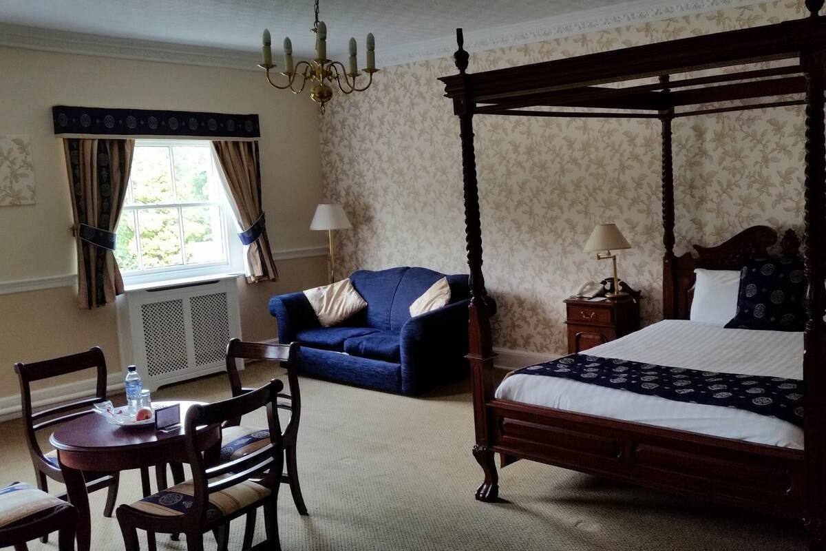 Una cama con dosel con cabecera de madera, un sofá azul, una mesa con sillas y una ventana con cortinas.