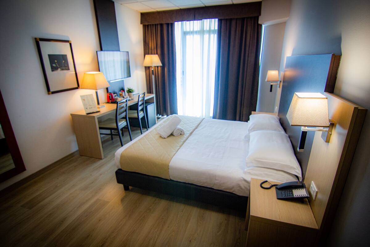 Quarto de hotel com cama, escrivaninha, cadeira, luminárias e telefone.