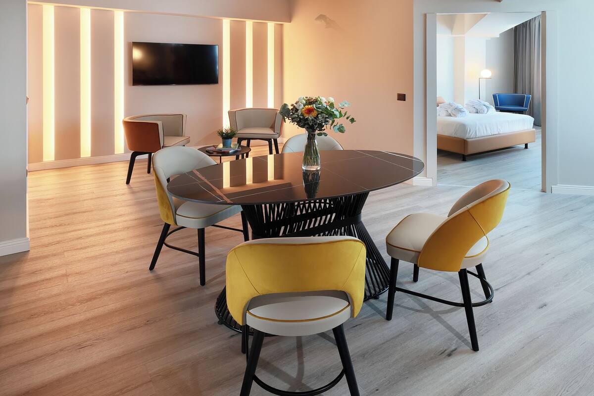 Quarto de hotel moderno com área de refeições, mesa redonda e cadeiras amarelas, televisão de tela plana fixada na parede e uma cama com uma cadeira azul no quarto vizinho.