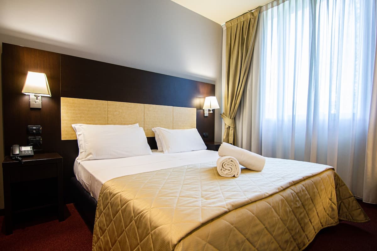 Quarto de hotel com uma cama grande, duas luminárias de cabeceira, uma mesa de cabeceira e uma janela com cortinas.