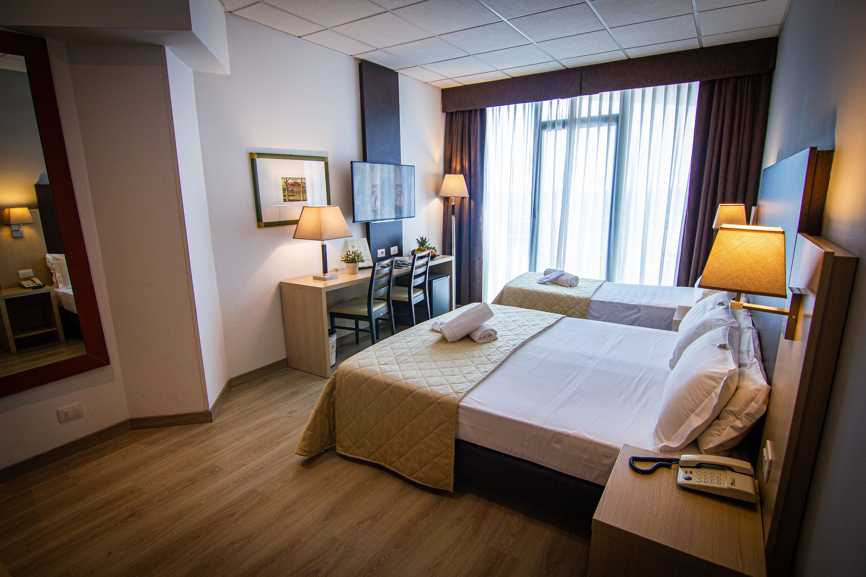 A hotel room with two beds, a desk with a chair, a telephone, and a window with curtains.