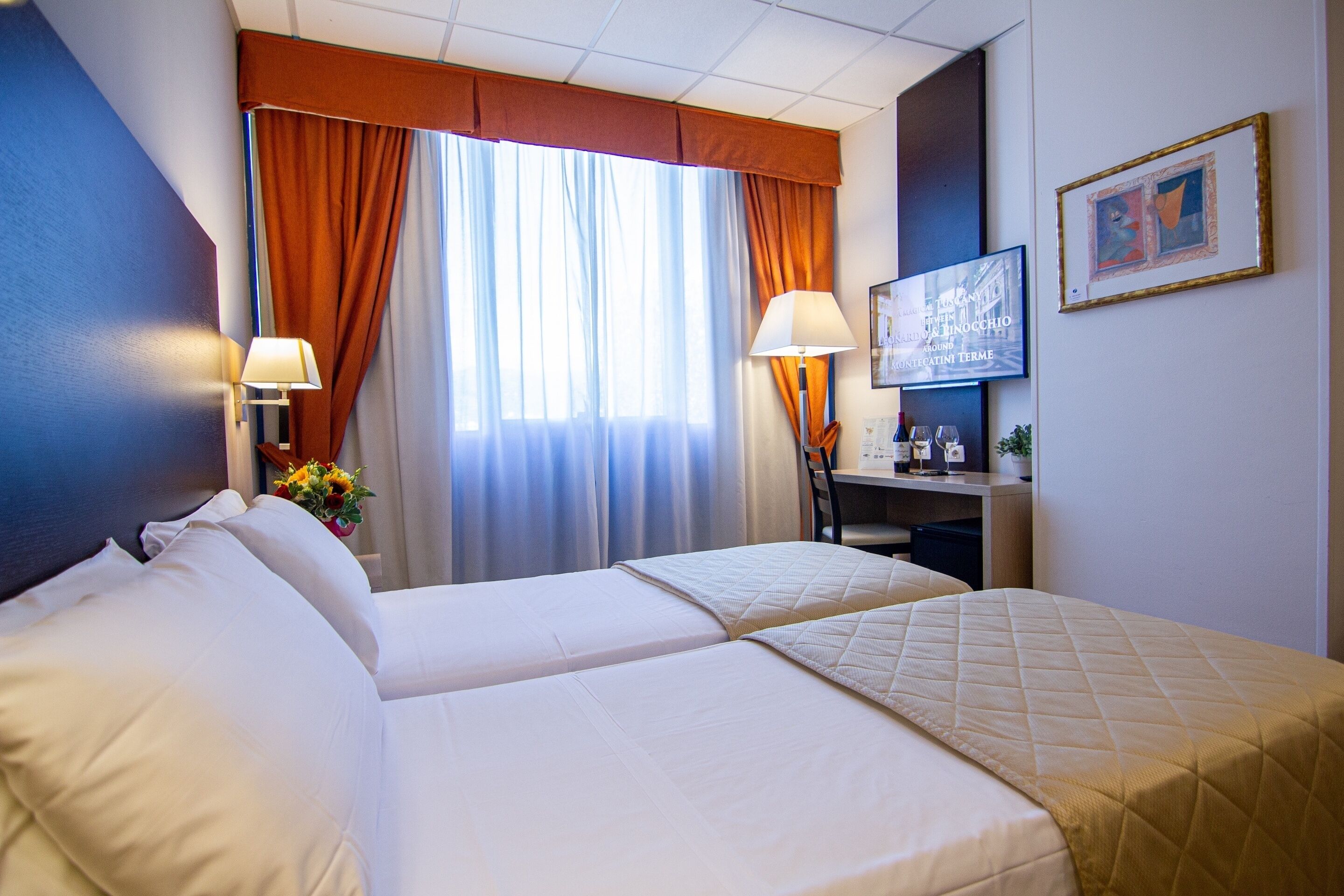A hotel room with two beds, a desk with a lamp, a television, and a framed picture on the wall.