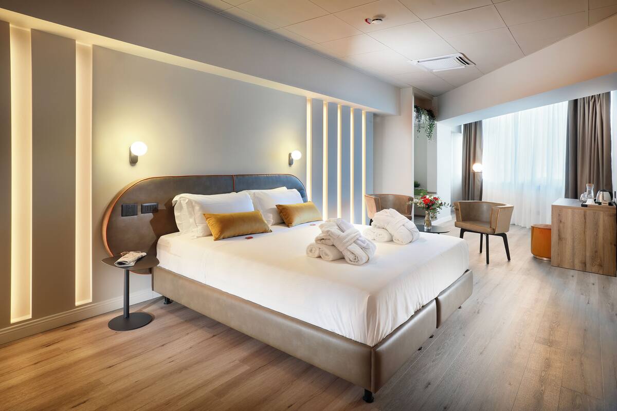 Quarto de hotel moderno com uma cama grande, mesas de cabeceira, escrivaninha e vista para o exterior.
