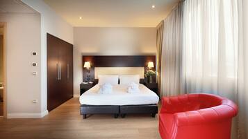 Habitación de hotel moderna con una cama grande, lámparas de noche, un sillón rojo y vistas al exterior a través de cortinas translúcidas.