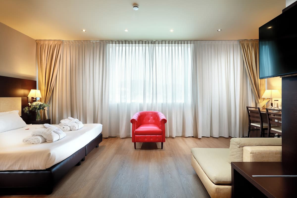 Quarto de hotel com cama, uma poltrona vermelha, sofá, televisão e área de refeições.